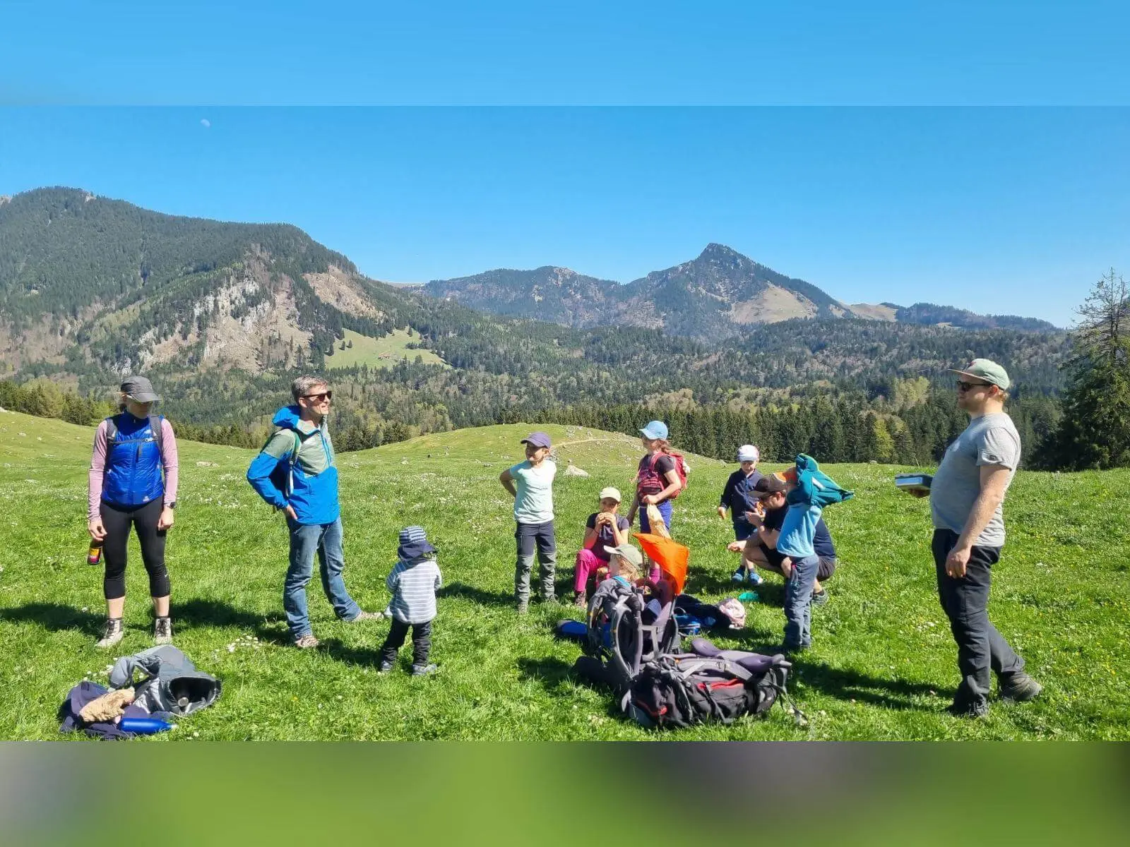 Familiengruppe mit Kindern macht Pause auf grüner Almwiese; Rucksäcke liegen am Boden, im Hintergrund Bergpanorama. | © DAV Markt Schwaben/Maria Wenisch