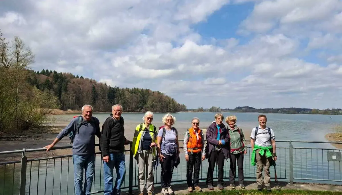 Gruppe älterer Wandernder steht am Ufer des Seehamer Sees vor Geländer und Wasser; im Hintergrund Uferwald und Wolkenhimmel. | © DAV Markt Schwaben/Rudolf Mandausch