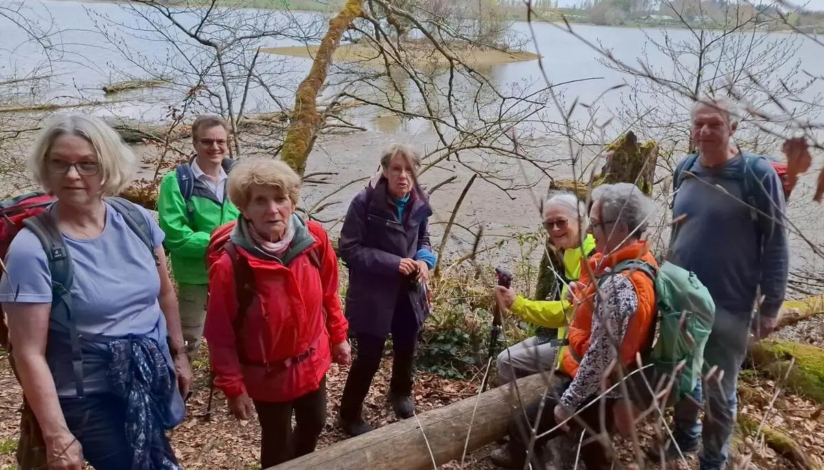 Wandergruppe steht auf schmalem Pfad im lichten Uferwald des Seehamer Sees; im Hintergrund Wasserfläche und gegenüberliegendes Ufer. | © DAV Markt Schwaben/Rudolf Mandausch