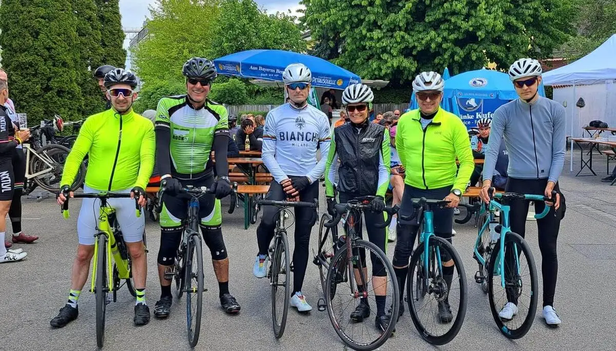 Sechs Radfahrende stehen mit Rennrädern nebeneinander auf einem Platz, im Hintergrund Biertische, Sonnenschirme und weitere Teilnehmende. | © DAV Markt Schwaben/Miron Thoms