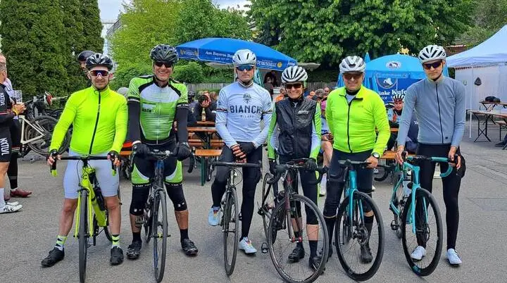 Sechs Radfahrende stehen mit Rennrädern nebeneinander auf einem Platz, im Hintergrund Biertische, Sonnenschirme und weitere Teilnehmende. | © DAV Markt Schwaben/Miron Thoms