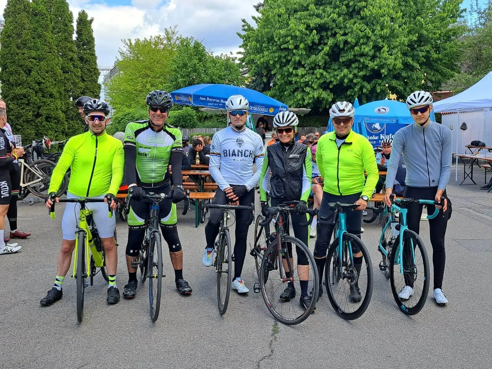 Sechs Radfahrende stehen mit Rennrädern nebeneinander auf einem Platz, im Hintergrund Biertische, Sonnenschirme und weitere Teilnehmende. | © DAV Markt Schwaben/Miron Thoms