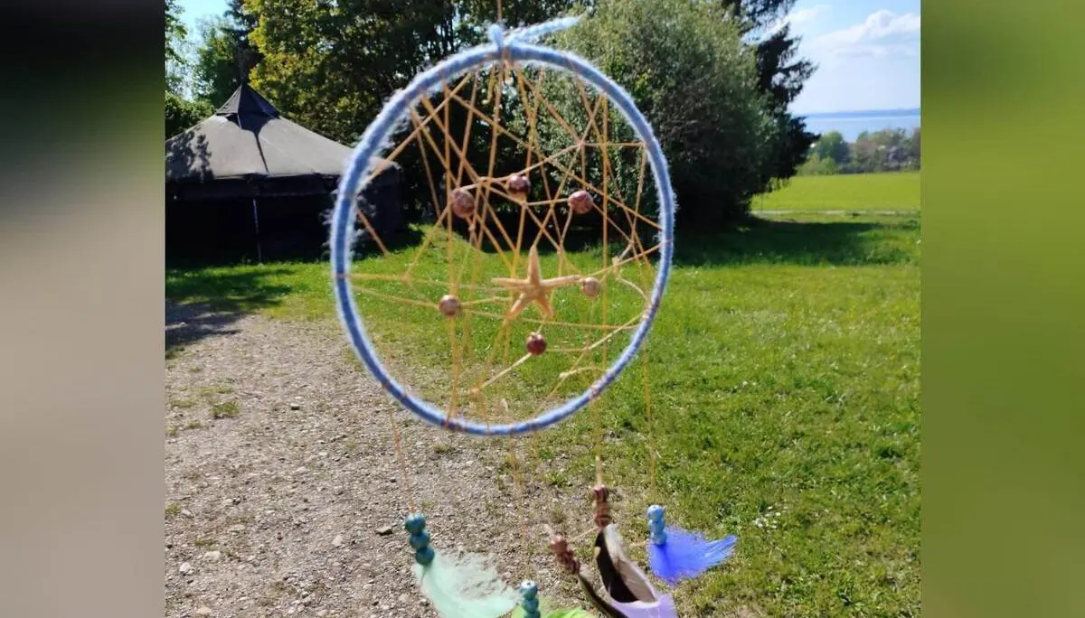 Selbst gebastelter Traumfänger mit Netz, Perlen und bunten Federn hängt im Vordergrund; dahinter Wiese, Bäume, ein Tipi und ferner See. | © DAV Markt Schwaben