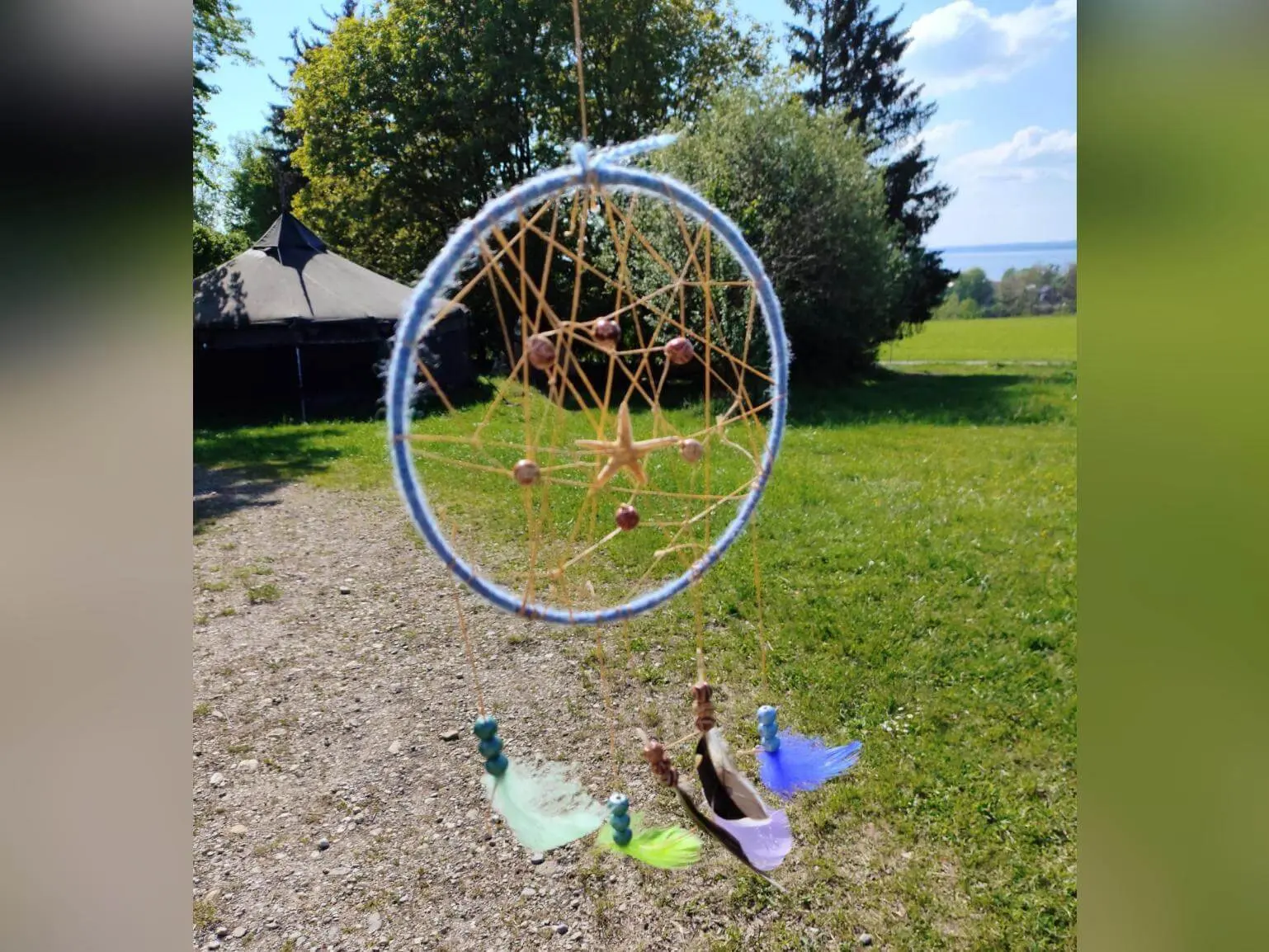 Selbst gebastelter Traumfänger mit Netz, Perlen und bunten Federn hängt im Vordergrund; dahinter Wiese, Bäume, ein Tipi und ferner See. | © DAV Markt Schwaben