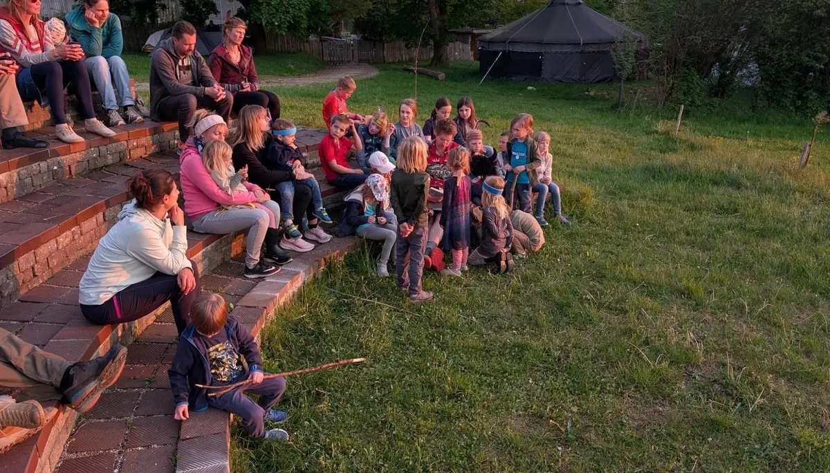 Kinder und Erwachsene sitzen auf Stufen im Freien und hören einer Person zu; im Hintergrund steht ein Tipi, warmes Abendlicht fällt auf die Wiese. | © DAV Markt Schwaben