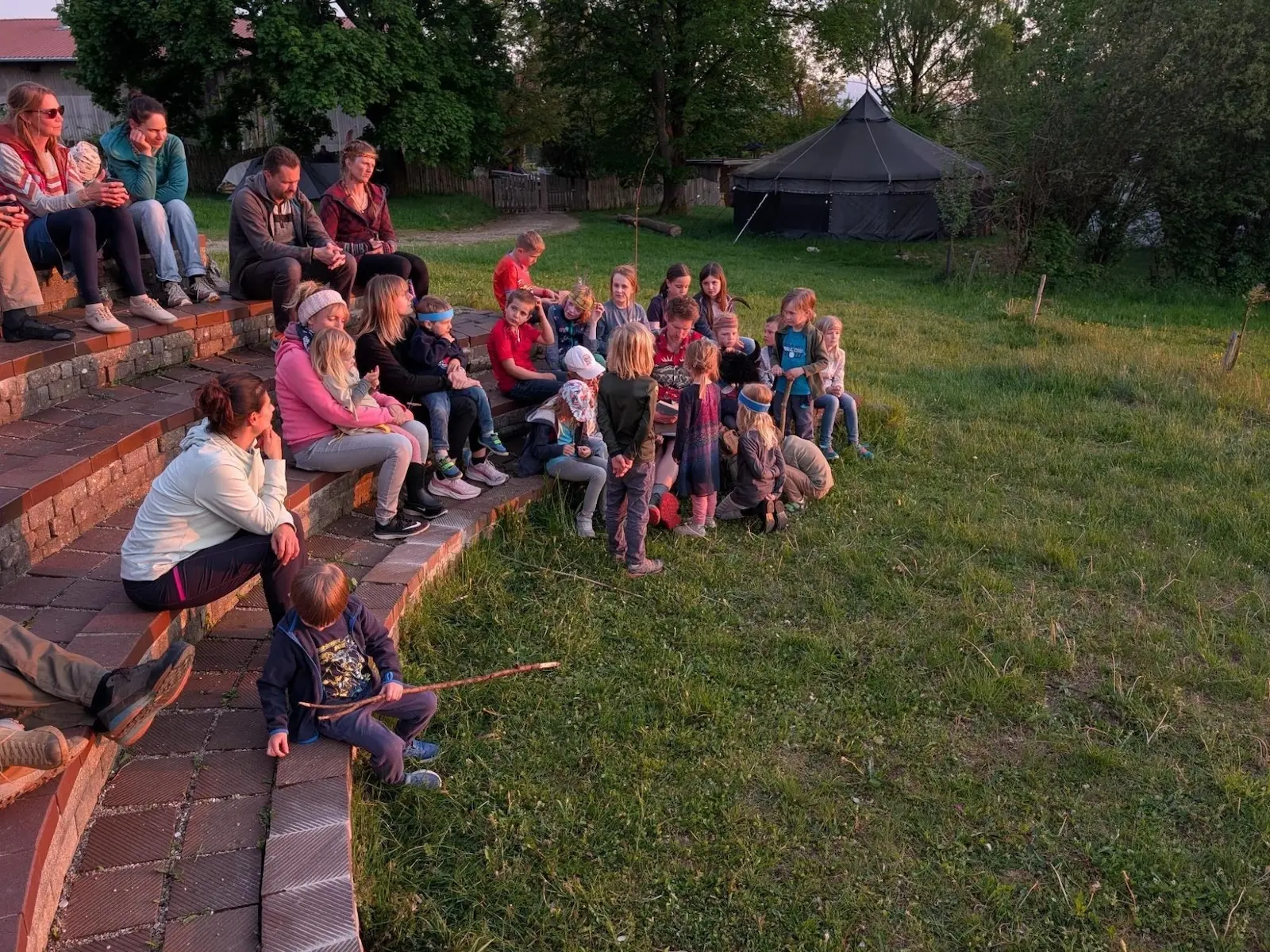 Kinder und Erwachsene sitzen auf Stufen im Freien und hören einer Person zu; im Hintergrund steht ein Tipi, warmes Abendlicht fällt auf die Wiese. | © DAV Markt Schwaben