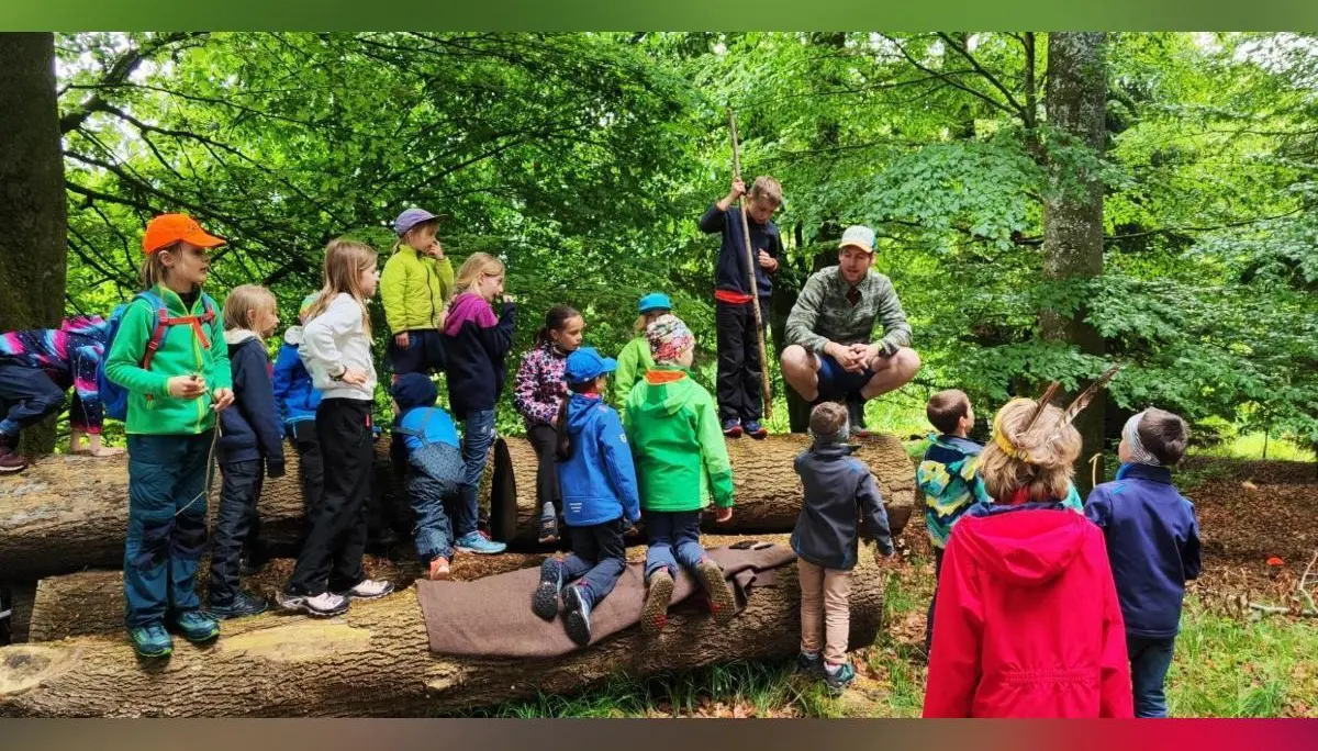 Kinder stehen und sitzen auf einem umgestürzten Baumstamm im Wald, während eine erwachsene Person Aufgaben erklärt; dichter grüner Wald umgibt die Szene. | © DAV Markt Schwaben