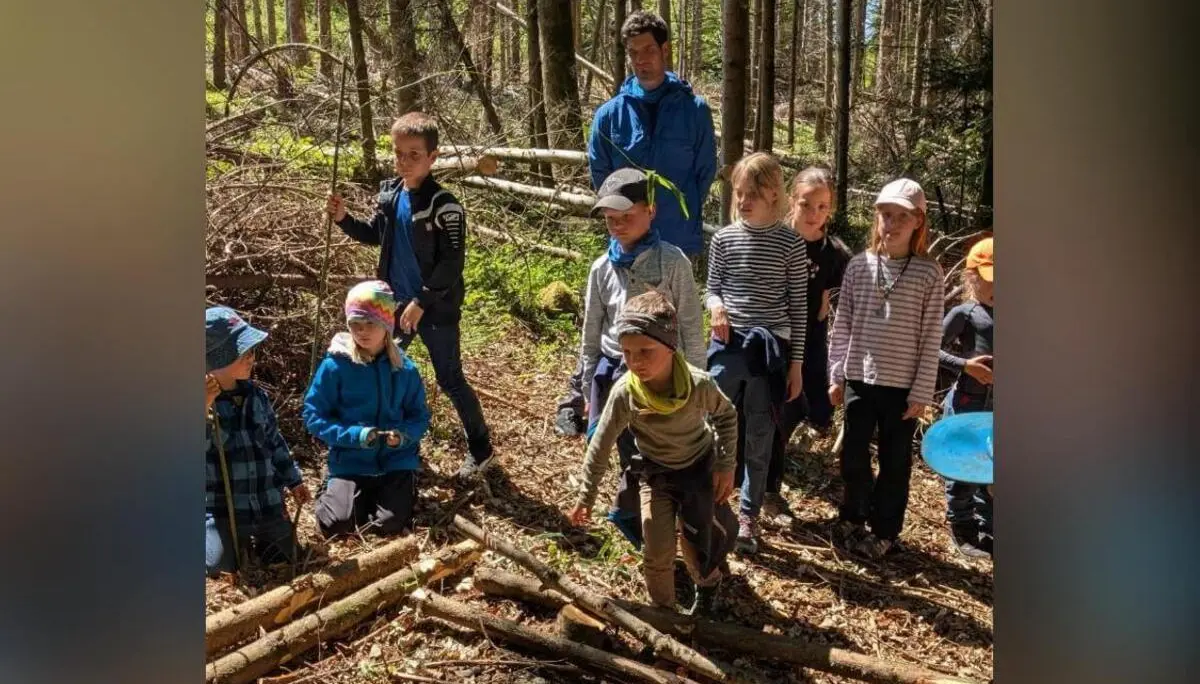 Mehrere Kinder stehen und knien zwischen Ästen und Stämmen im Wald, während eine erwachsene Person dahinter beobachtet; ein Kind balanciert über Holzstücke. | © DAV Markt Schwaben