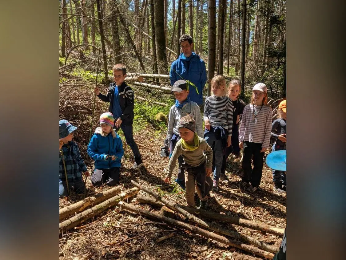 Mehrere Kinder stehen und knien zwischen Ästen und Stämmen im Wald, während eine erwachsene Person dahinter beobachtet; ein Kind balanciert über Holzstücke. | © DAV Markt Schwaben