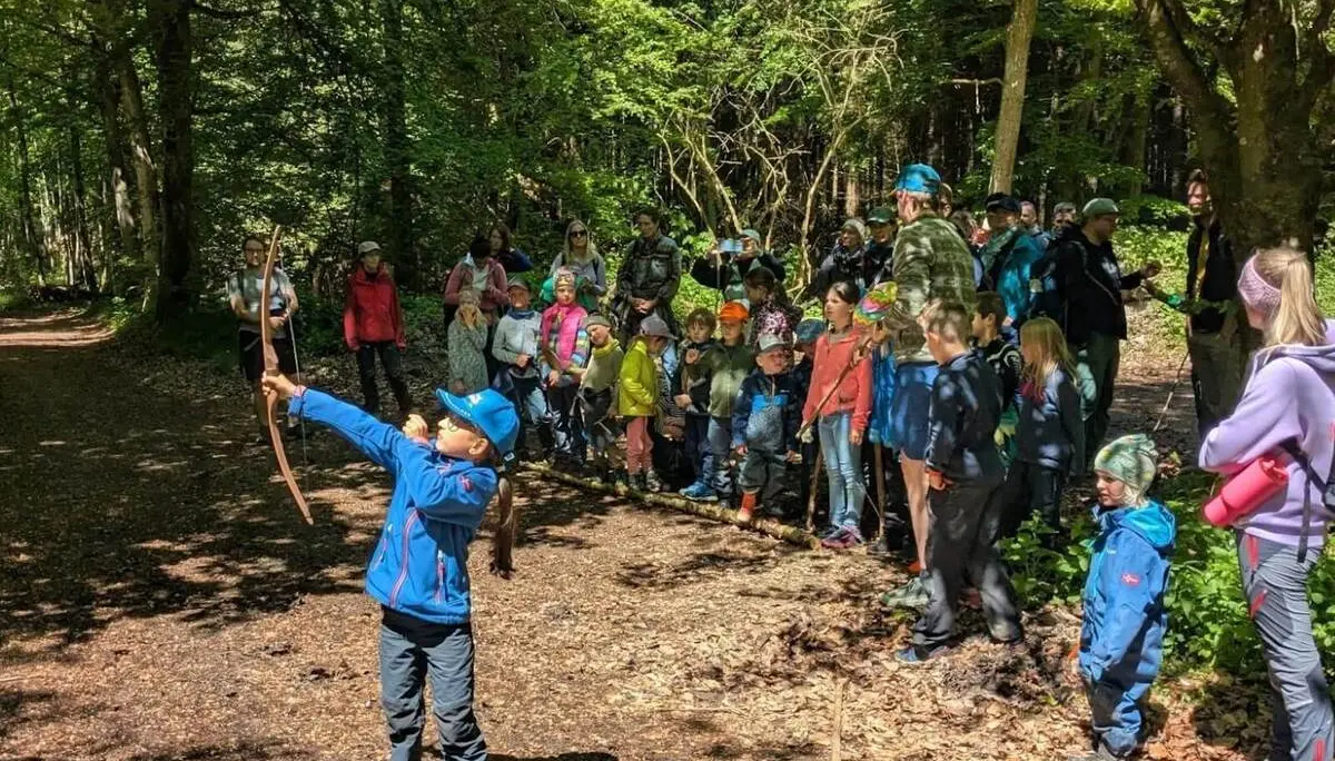 Kind spannt einen Bogen und zielt im Wald, während eine große Gruppe aus Kindern und Erwachsenen dahinter steht und zusieht. | © DAV Markt Schwaben