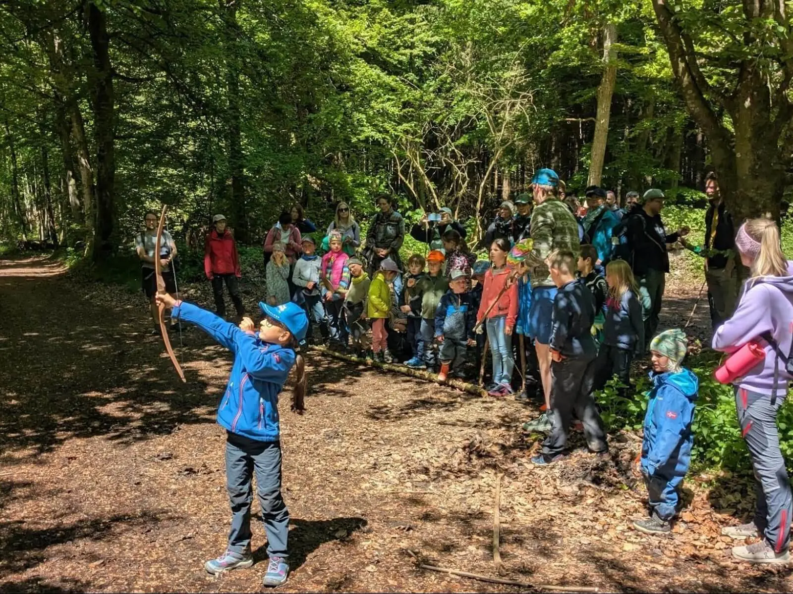 Kind spannt einen Bogen und zielt im Wald, während eine große Gruppe aus Kindern und Erwachsenen dahinter steht und zusieht. | © DAV Markt Schwaben