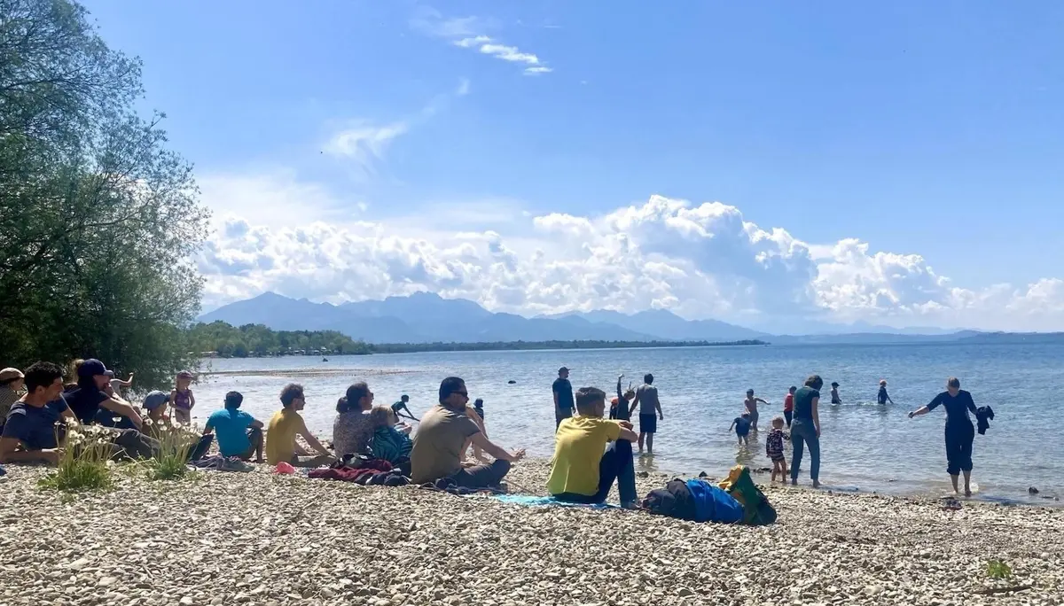 Kinder und Erwachsene sitzen und stehen am steinigen Ufer des Chiemsees, einige waten im Wasser; im Hintergrund Bergkette unter blauem Himmel. | © DAV Markt Schwaben