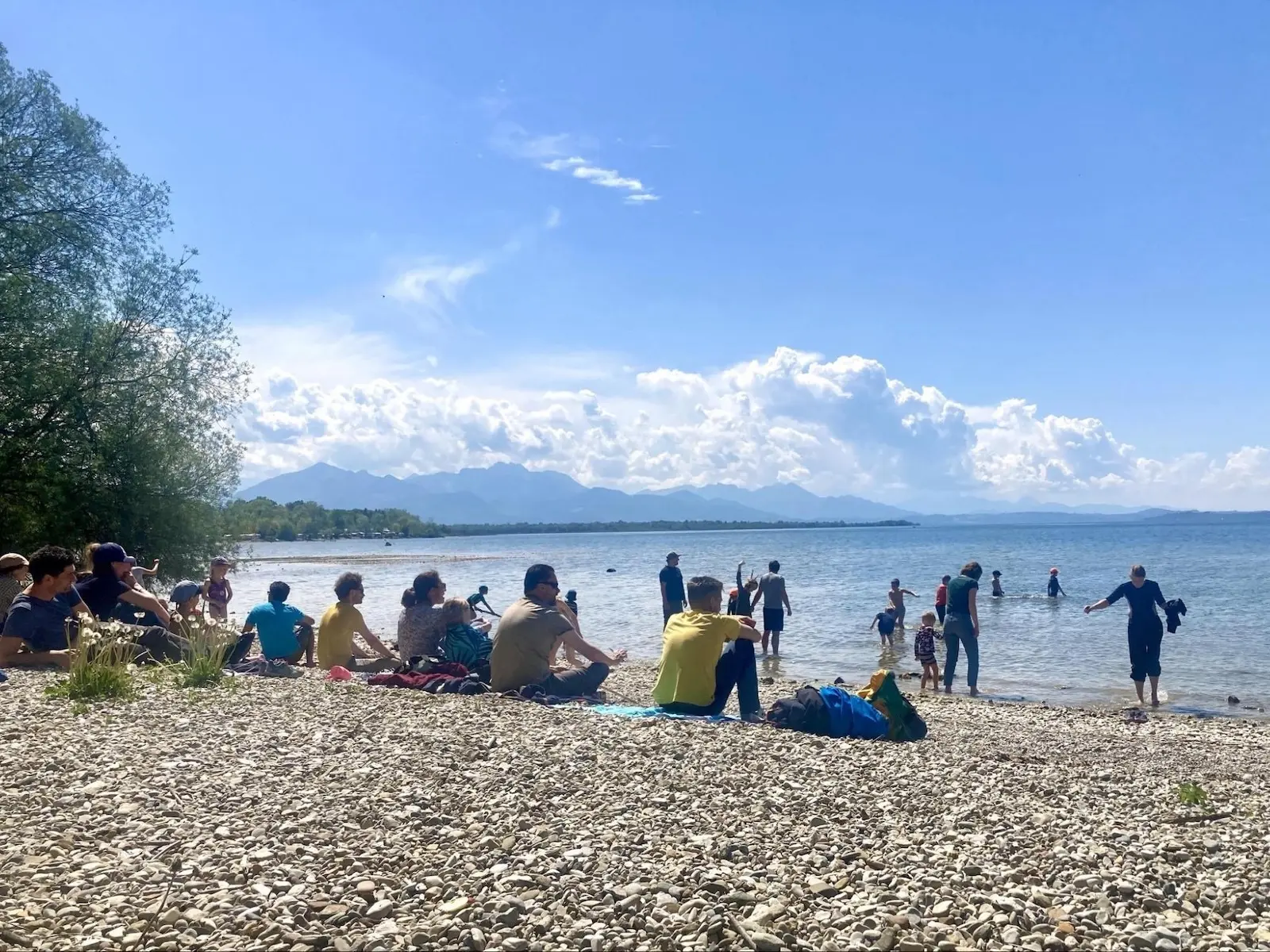 Kinder und Erwachsene sitzen und stehen am steinigen Ufer des Chiemsees, einige waten im Wasser; im Hintergrund Bergkette unter blauem Himmel. | © DAV Markt Schwaben