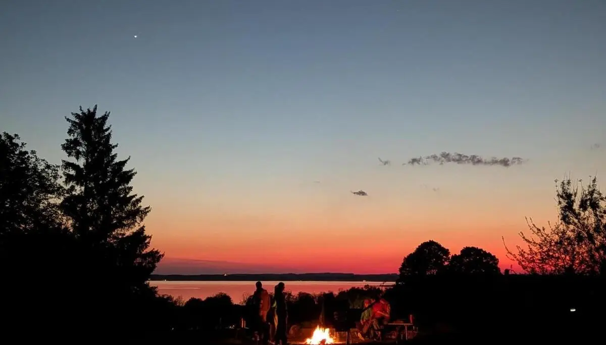 Mehrere Personen stehen und sitzen um ein Lagerfeuer am Ufer; im Hintergrund liegt der Chiemsee, darüber ein rot-blauer Abendhimmel mit ersten Sternen. | © DAV Markt Schwaben