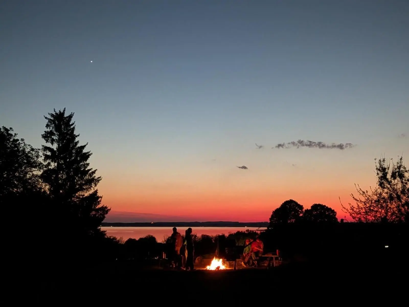 Mehrere Personen stehen und sitzen um ein Lagerfeuer am Ufer; im Hintergrund liegt der Chiemsee, darüber ein rot-blauer Abendhimmel mit ersten Sternen. | © DAV Markt Schwaben