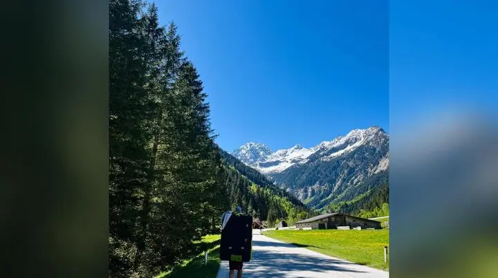 Person trägt Crashpad auf einer Straße im Zillertal, umgeben von Wald, Wiesen, Hütte und schneebedeckten Bergen unter blauem Himmel. | © DAV Markt Schwaben