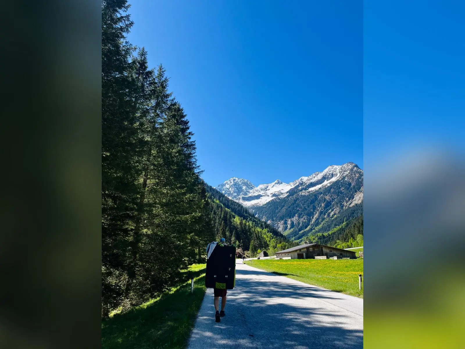Person trägt Crashpad auf einer Straße im Zillertal, umgeben von Wald, Wiesen, Hütte und schneebedeckten Bergen unter blauem Himmel. | © DAV Markt Schwaben