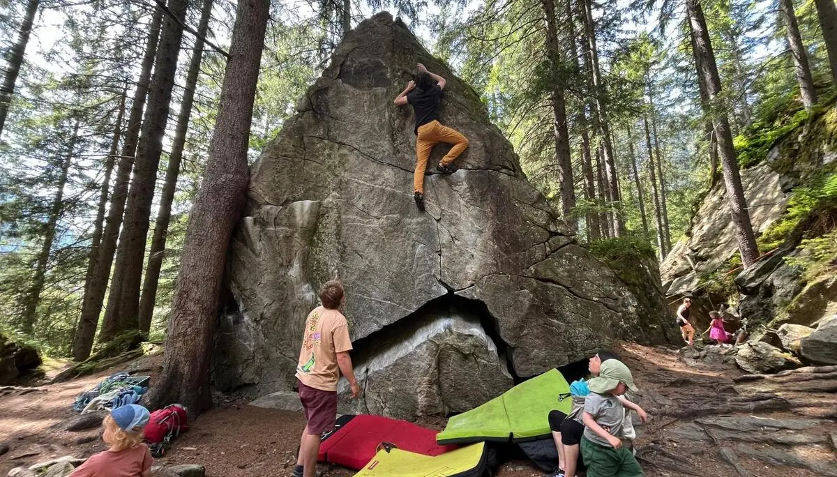 Mehrere Personen bouldern an einem großen Fels im Wald, Crashpads liegen bereit; Kinder spielen zwischen Felsen und Bäumen. | © DAV Markt Schwaben