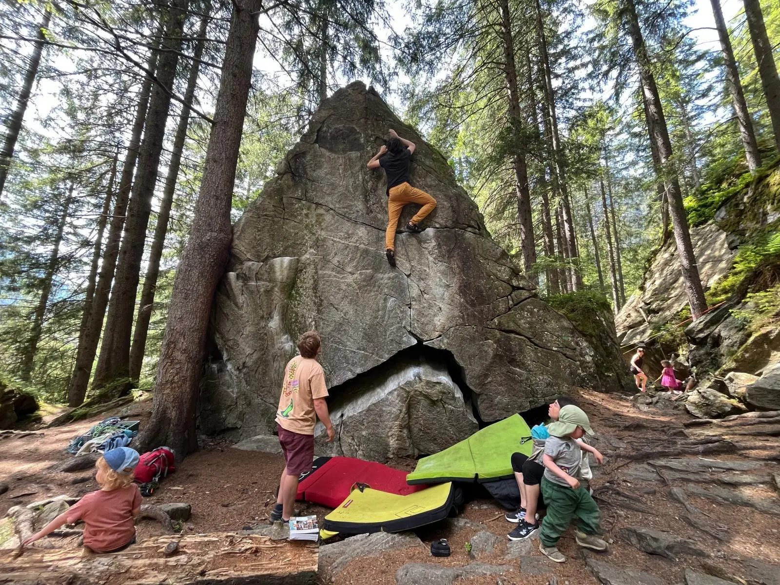 Mehrere Personen bouldern an einem großen Fels im Wald, Crashpads liegen bereit; Kinder spielen zwischen Felsen und Bäumen. | © DAV Markt Schwaben