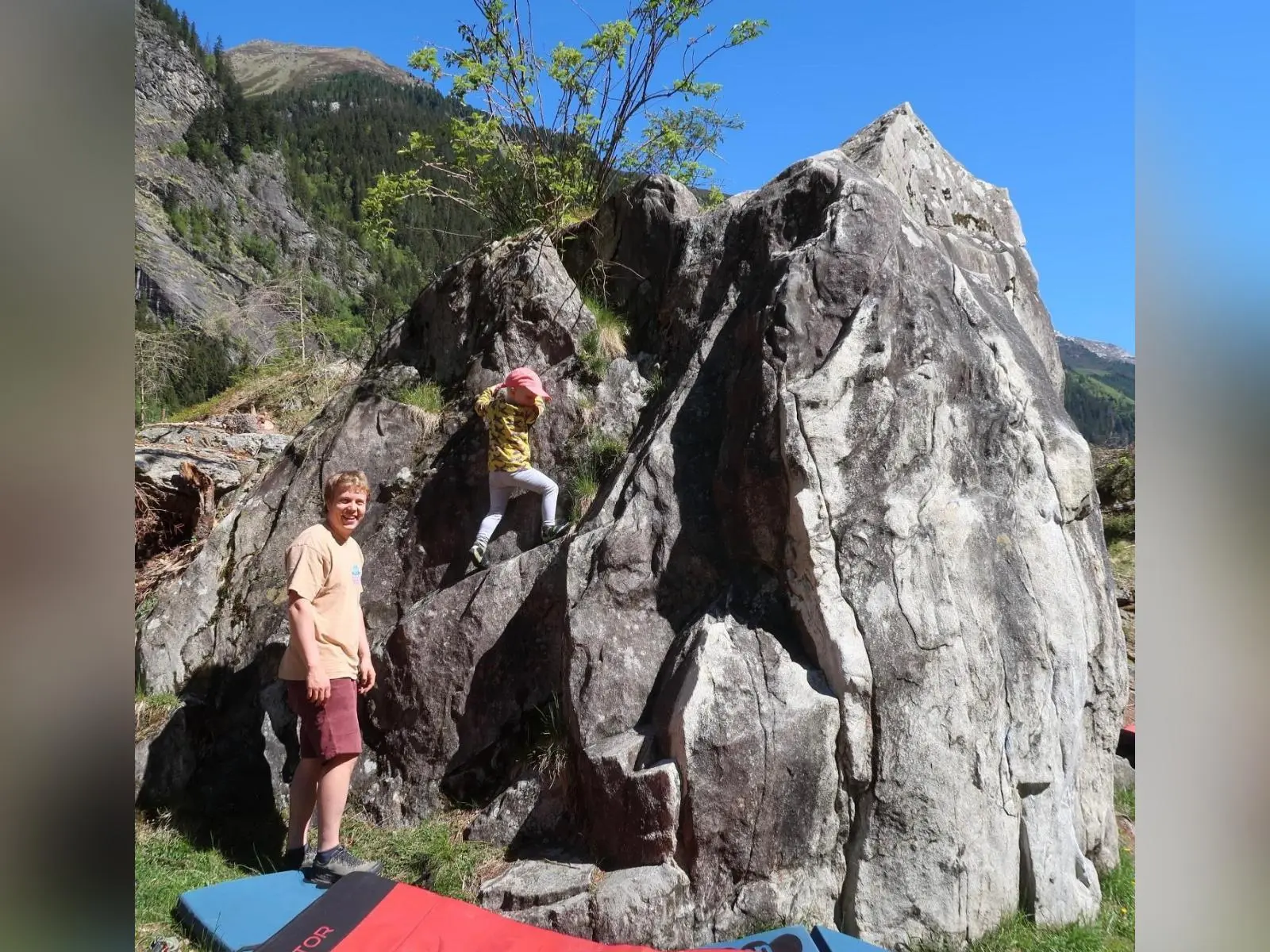 Kind klettert an Felsblock, Person steht daneben auf Crashpads; dahinter Berge und blauer Himmel. | © DAV Markt Schwaben
