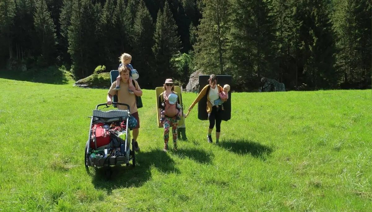 Mehrere Personen mit Kindern und Ausrüstung gehen über eine Wiese im Zillertal, tragen Crashpads und schieben einen Wagen vor Waldkulisse. | © DAV Markt Schwaben