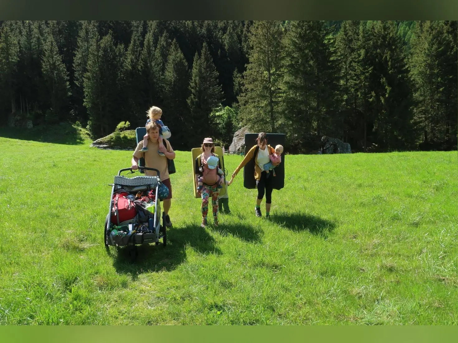 Mehrere Personen mit Kindern und Ausrüstung gehen über eine Wiese im Zillertal, tragen Crashpads und schieben einen Wagen vor Waldkulisse. | © DAV Markt Schwaben