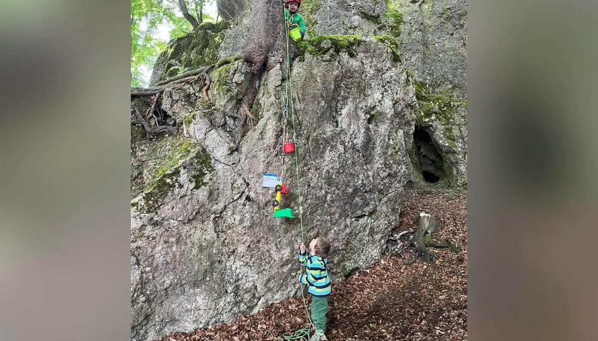 Zwei Kinder spielen am Kalkfelsen im Wald; eines steht oben mit Helm, das andere zieht unten ein Spielzeug am Seil hoch. | © DAV Markt Schwaben / Lara Wolff