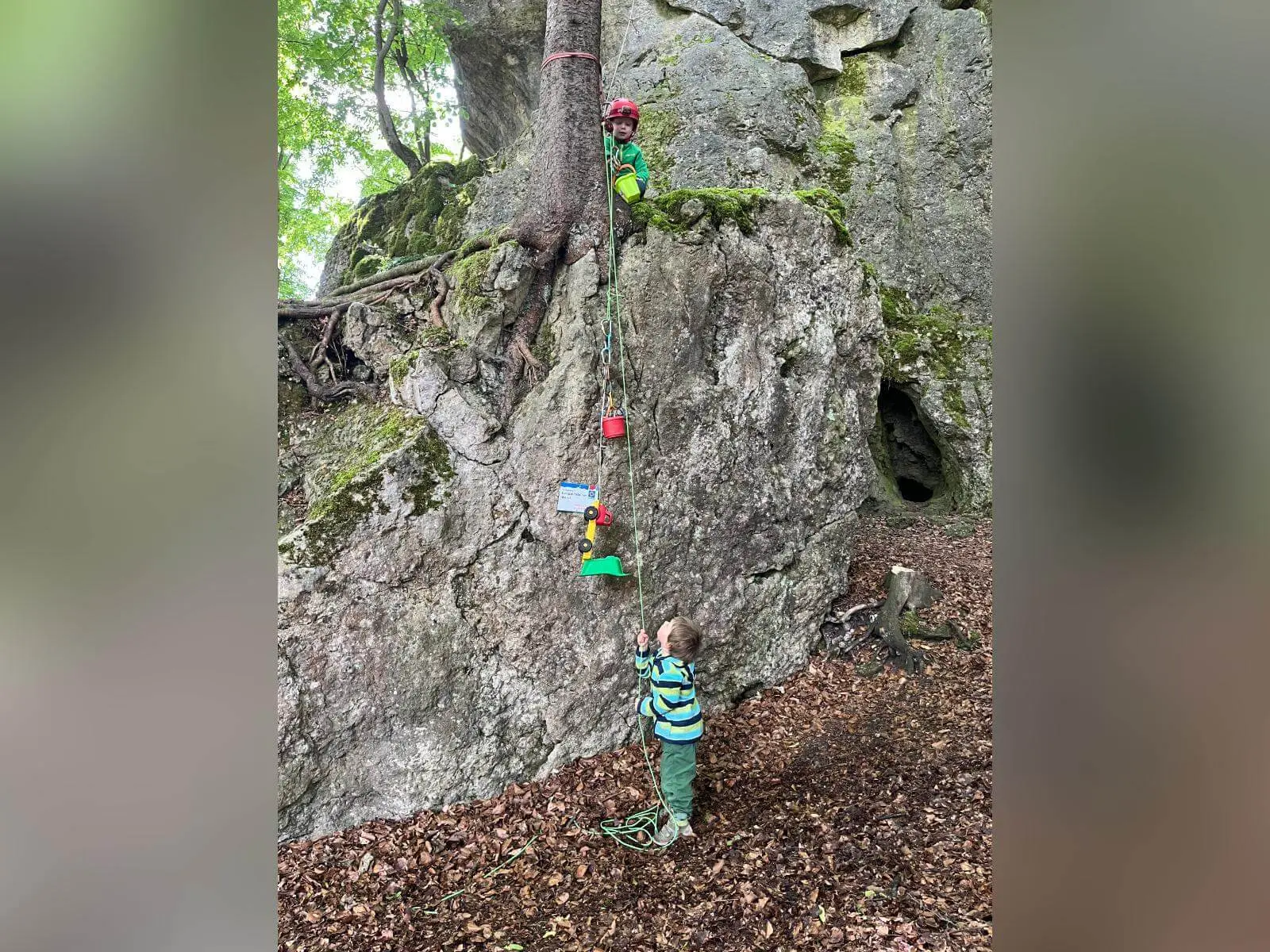 Zwei Kinder spielen am Kalkfelsen im Wald; eines steht oben mit Helm, das andere zieht unten ein Spielzeug am Seil hoch. | © DAV Markt Schwaben / Lara Wolff