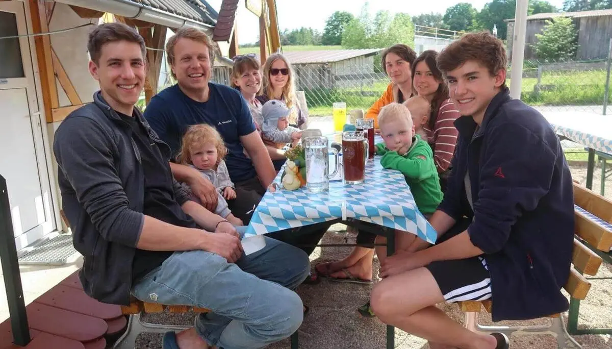 Gruppe sitzt an einem Biertisch im Freien, mit Getränken und Kindern; im Hintergrund sind Wiese, Gebäude und Sonnenschirm. | © DAV Markt Schwaben / Lara Wolff