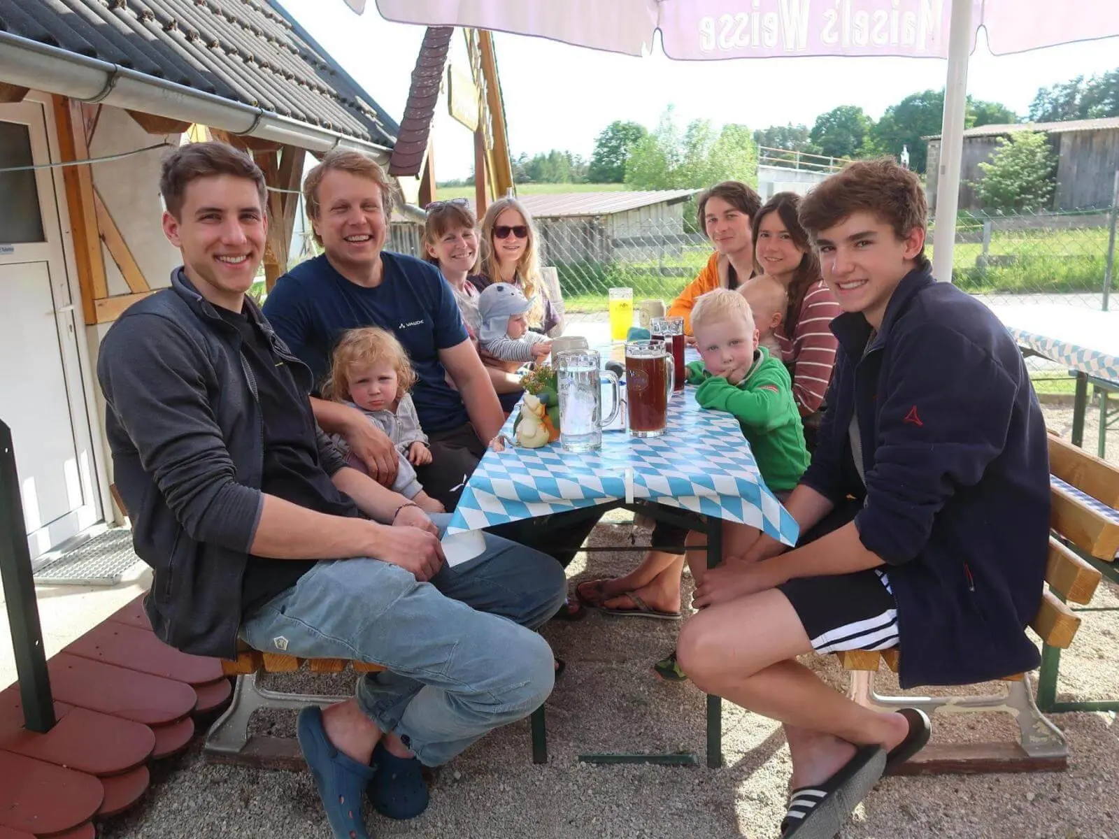 Gruppe sitzt an einem Biertisch im Freien, mit Getränken und Kindern; im Hintergrund sind Wiese, Gebäude und Sonnenschirm. | © DAV Markt Schwaben / Lara Wolff