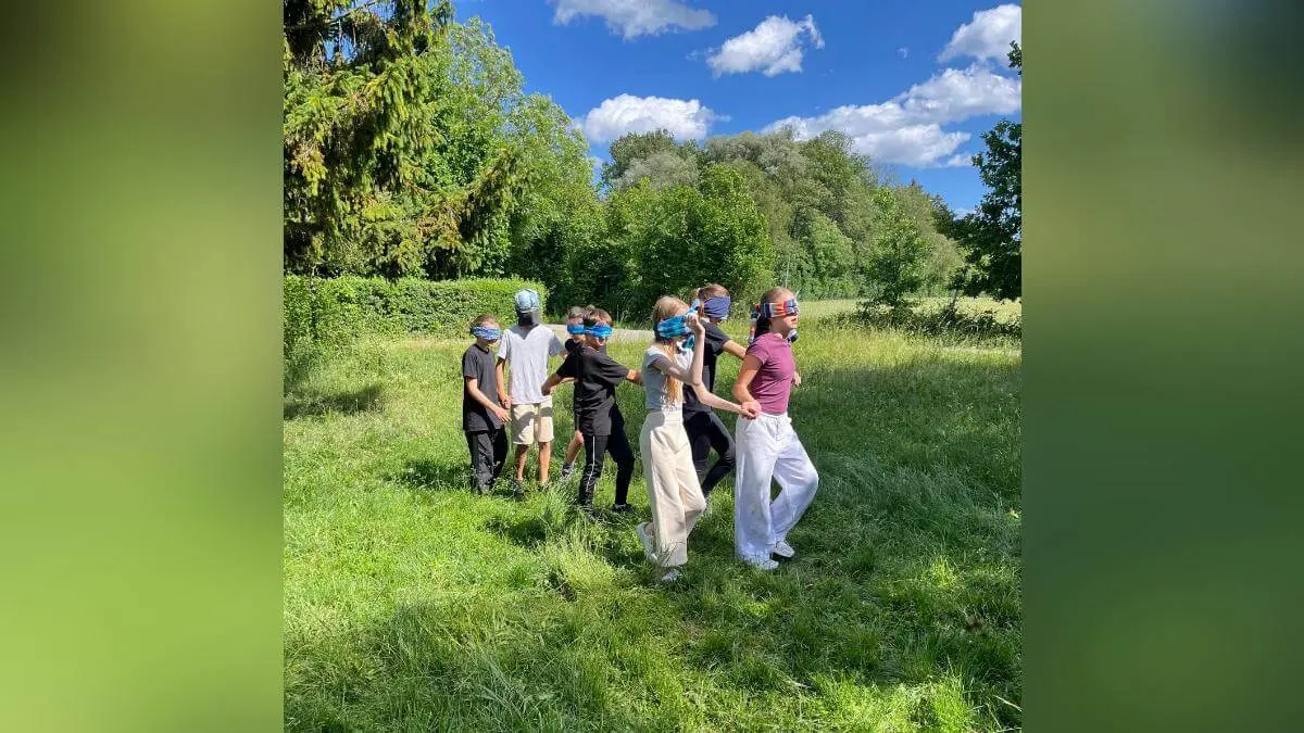 Mehrere Kinder stehen in einer Reihe auf einer Wiese und spielen gemeinsam ein Bewegungsspiel im Freien mit verbundenen Augen. | © DAV Markt Schwaben /  Wiebke Rauschenbach