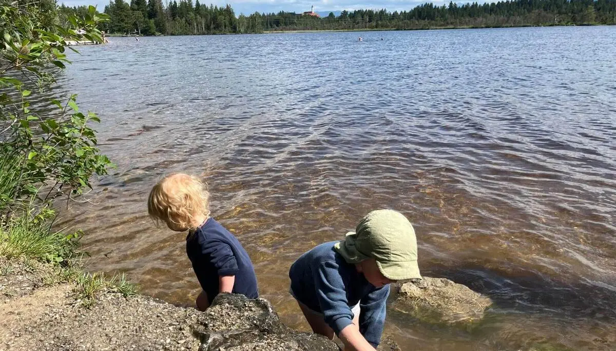 Zwei Kinder spielen am Ufer eines Sees und greifen ins flache Wasser, während im Hintergrund weitere Personen im See schwimmen. | © DAV Markt Schwaben / Lara Wolff