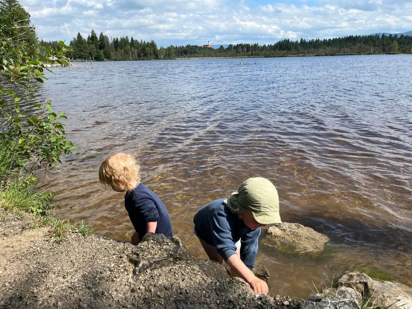 Zwei Kinder spielen am Ufer eines Sees und greifen ins flache Wasser, während im Hintergrund weitere Personen im See schwimmen. | © DAV Markt Schwaben / Lara Wolff
