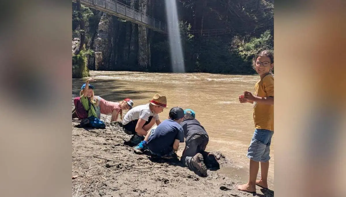 Mehrere Kinder knien am schlammigen Ufer eines braunen Flusses, spielen im Matsch; im Hintergrund eine Felswand mit Hängebrücke und Sonnenstrahl. | © DAV Markt Schwaben / Silke Titze