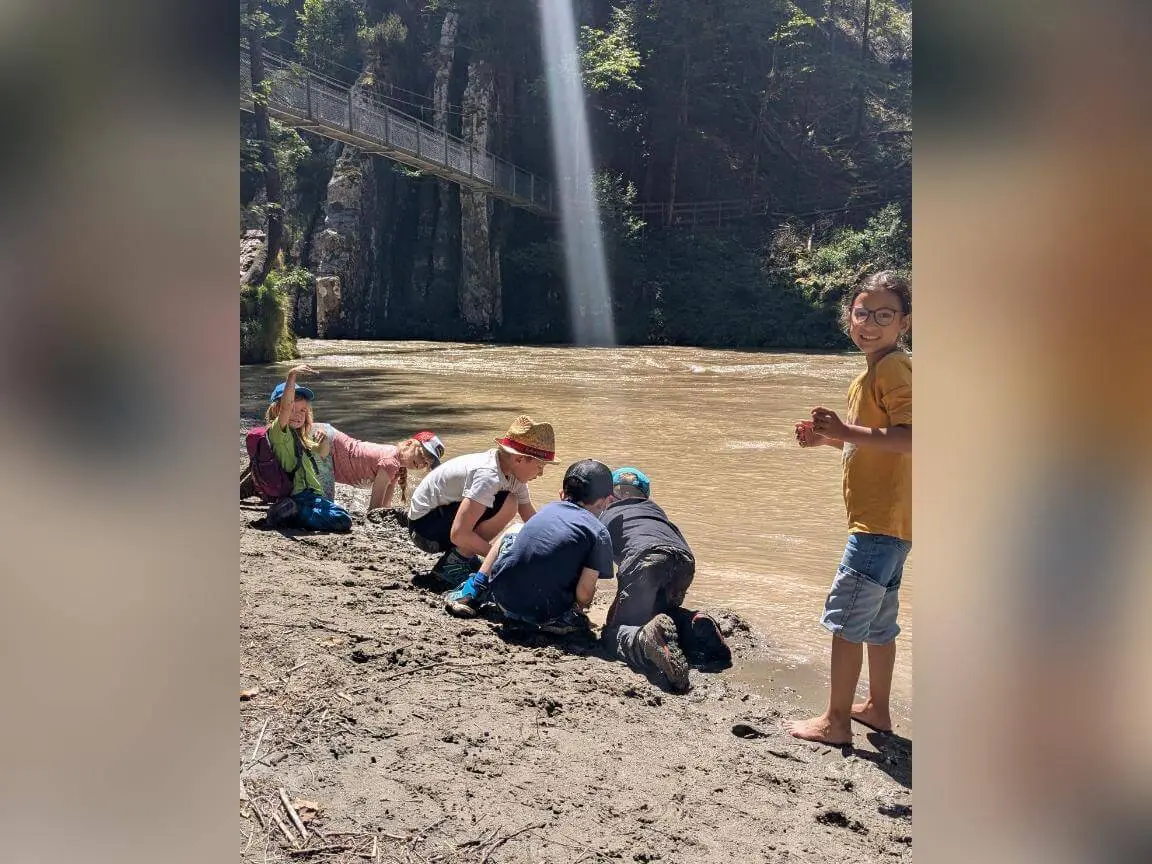 Mehrere Kinder knien am schlammigen Ufer eines braunen Flusses, spielen im Matsch; im Hintergrund eine Felswand mit Hängebrücke und Sonnenstrahl. | © DAV Markt Schwaben / Silke Titze