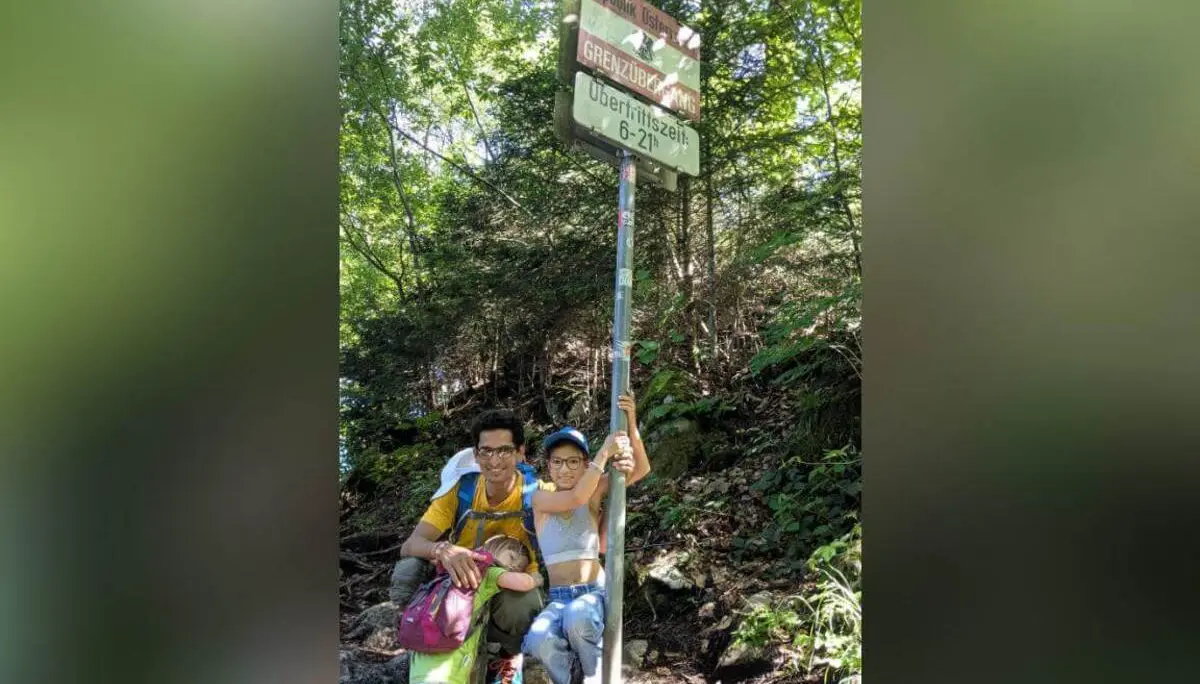 Zwei Personen stehen an einem Schild mit „Grenzübergang“ im Wald, umgeben von Bäumen und Felsen entlang eines Wanderwegs. | © DAV Markt Schwaben / Silke Titze