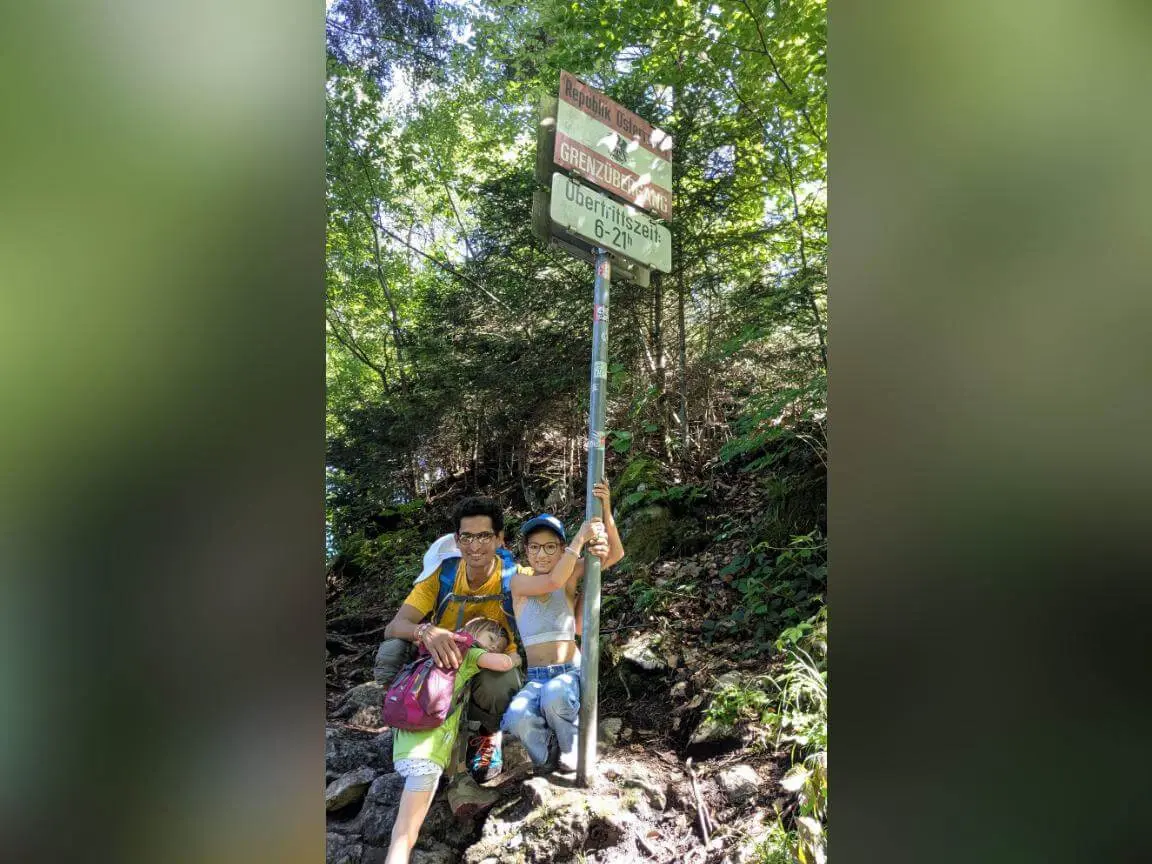Zwei Personen stehen an einem Schild mit „Grenzübergang“ im Wald, umgeben von Bäumen und Felsen entlang eines Wanderwegs. | © DAV Markt Schwaben / Silke Titze