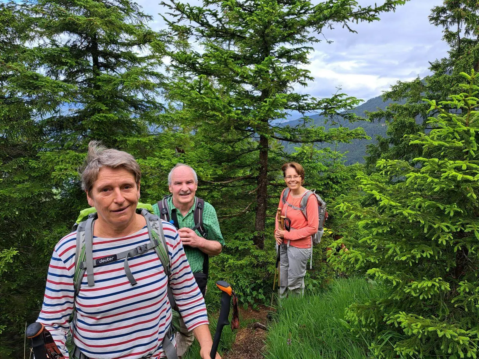 Drei Personen mit Rucksäcken und Wanderstöcken stehen auf einem schmalen Waldpfad zwischen dichten Fichten am Berghang. | © DAV Markt Schwaben / Tanja Döhler