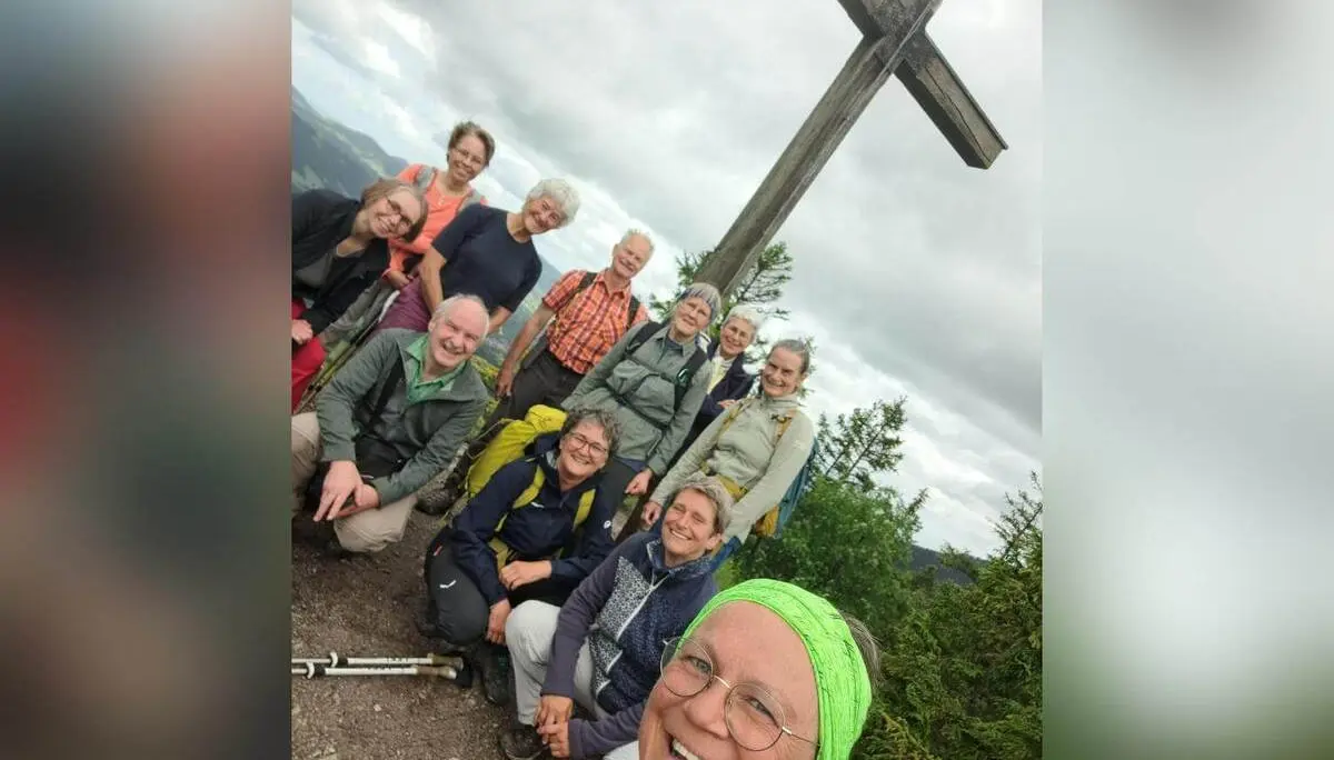 Zehn Wandernde posieren vor einem großen Holzgipfelkreuz auf einem Berggipfel; im Hintergrund ziehen dichte Wolken auf. | © DAV Markt Schwaben / Tanja Döhler