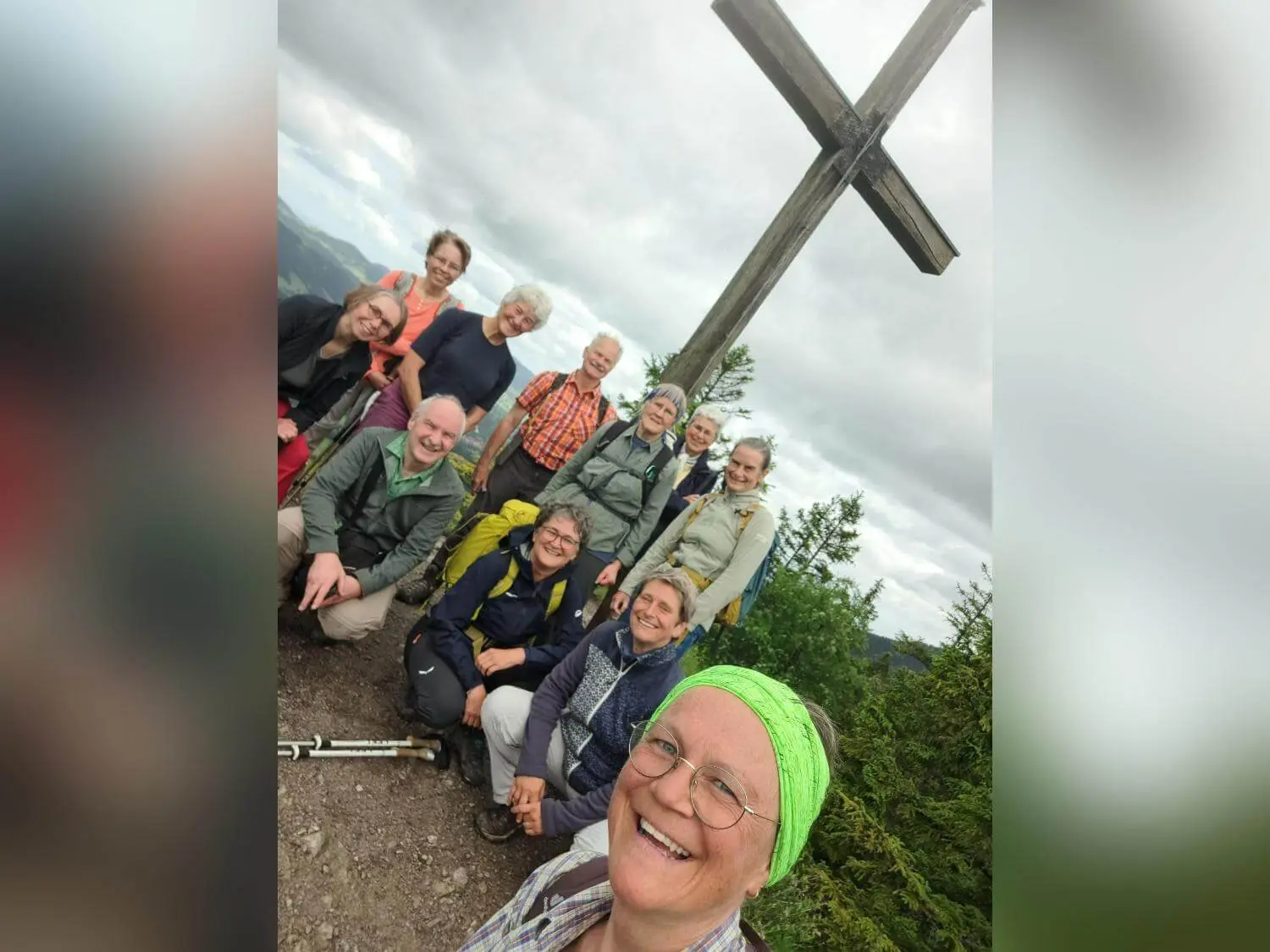 Zehn Wandernde posieren vor einem großen Holzgipfelkreuz auf einem Berggipfel; im Hintergrund ziehen dichte Wolken auf. | © DAV Markt Schwaben / Tanja Döhler