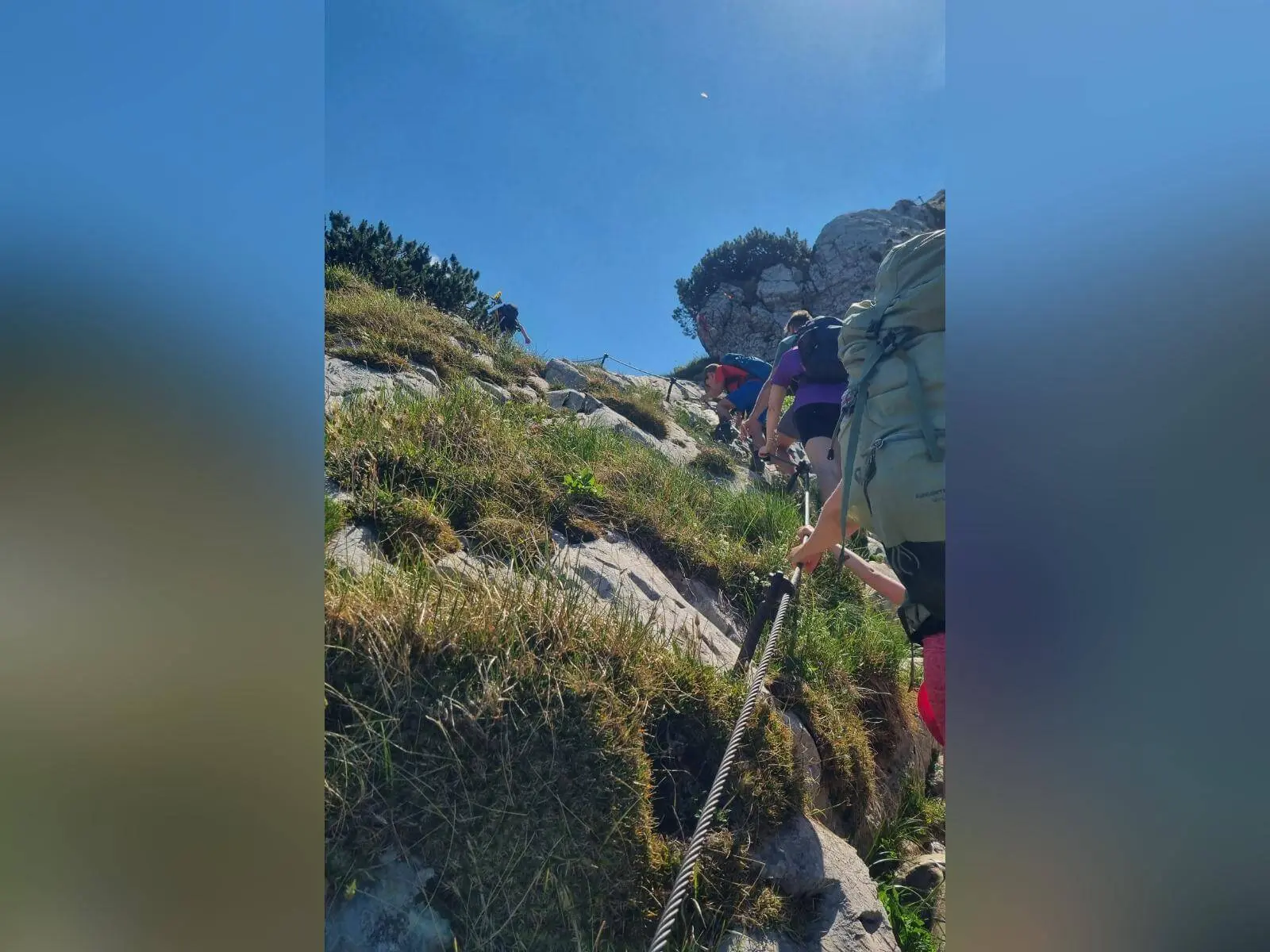 Mehrere Wandernde steigen an einem steilen, seilgesicherten Fels- und Grashang bergauf; über ihnen spannt sich ein wolkenloser Himmel. | © DAV Markt Schwaben