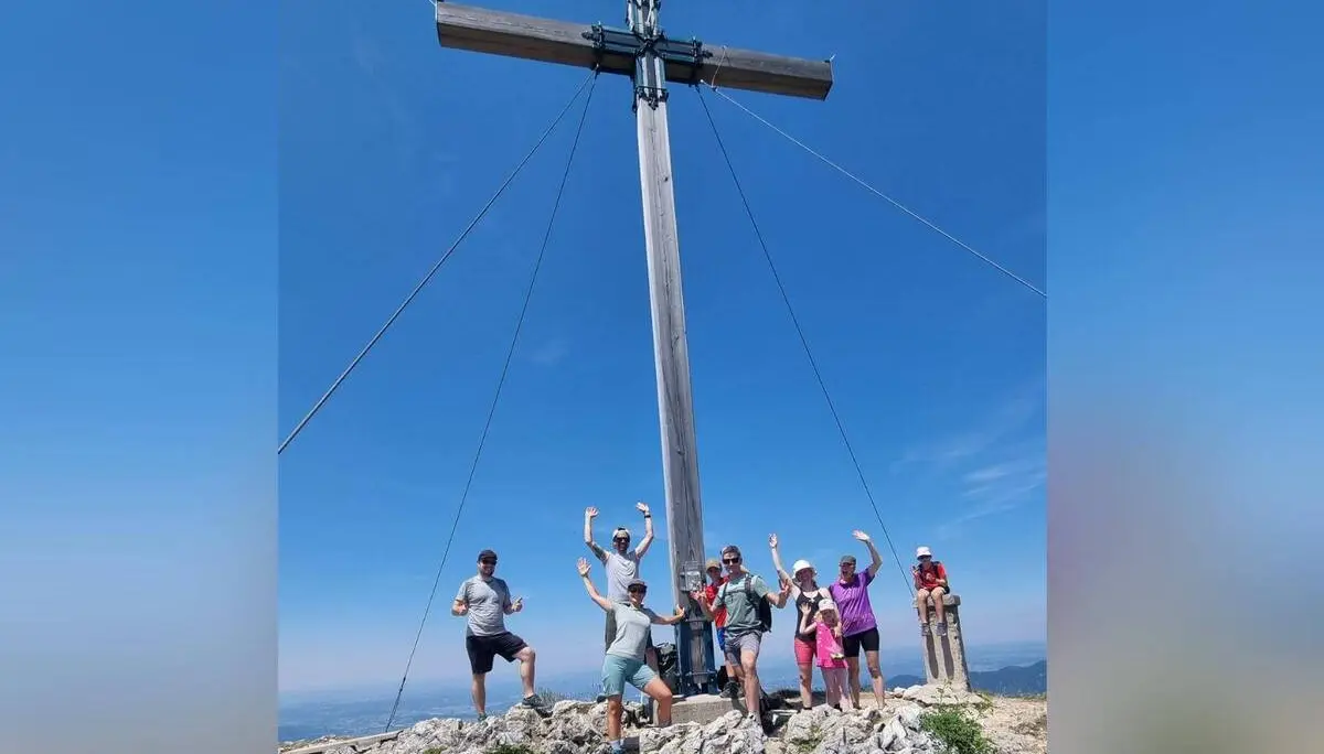 Wandergruppe steht auf felsigem Gipfelgelände unter einem großen Holzkreuz der Benediktenwand; der Himmel ist wolkenlos blau. | © DAV Markt Schwaben