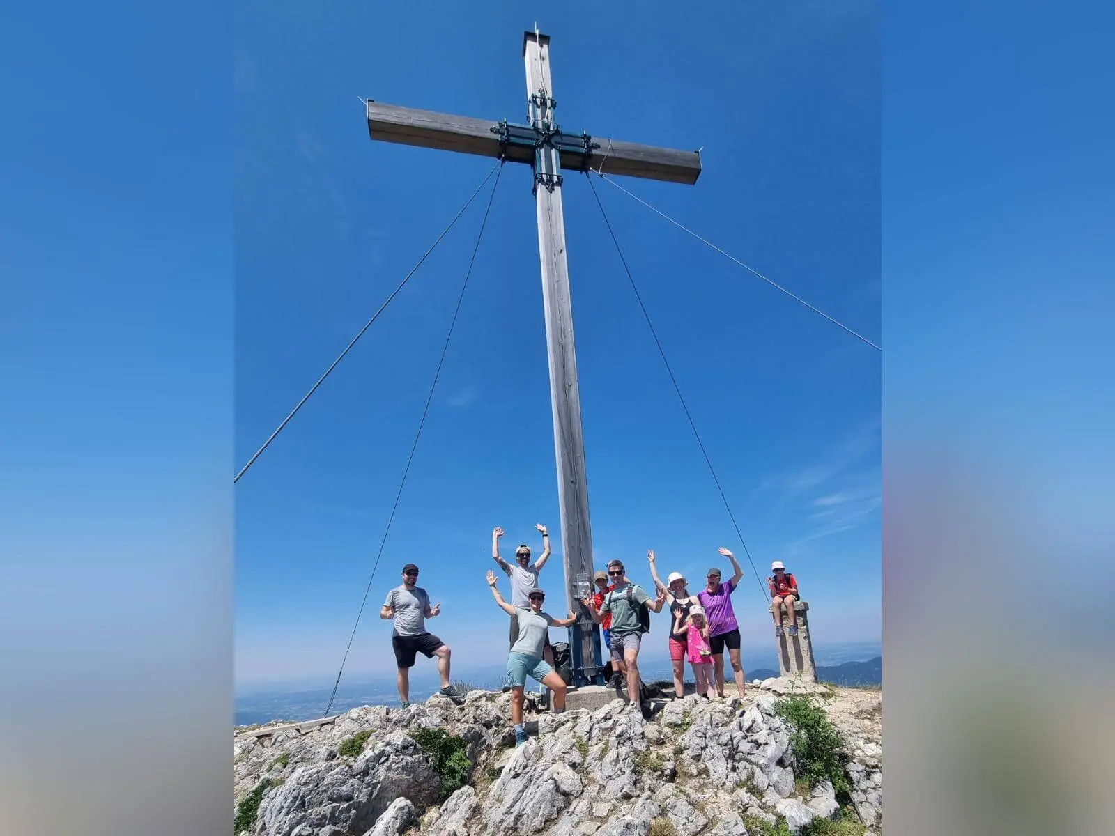 Wandergruppe steht auf felsigem Gipfelgelände unter einem großen Holzkreuz der Benediktenwand; der Himmel ist wolkenlos blau. | © DAV Markt Schwaben
