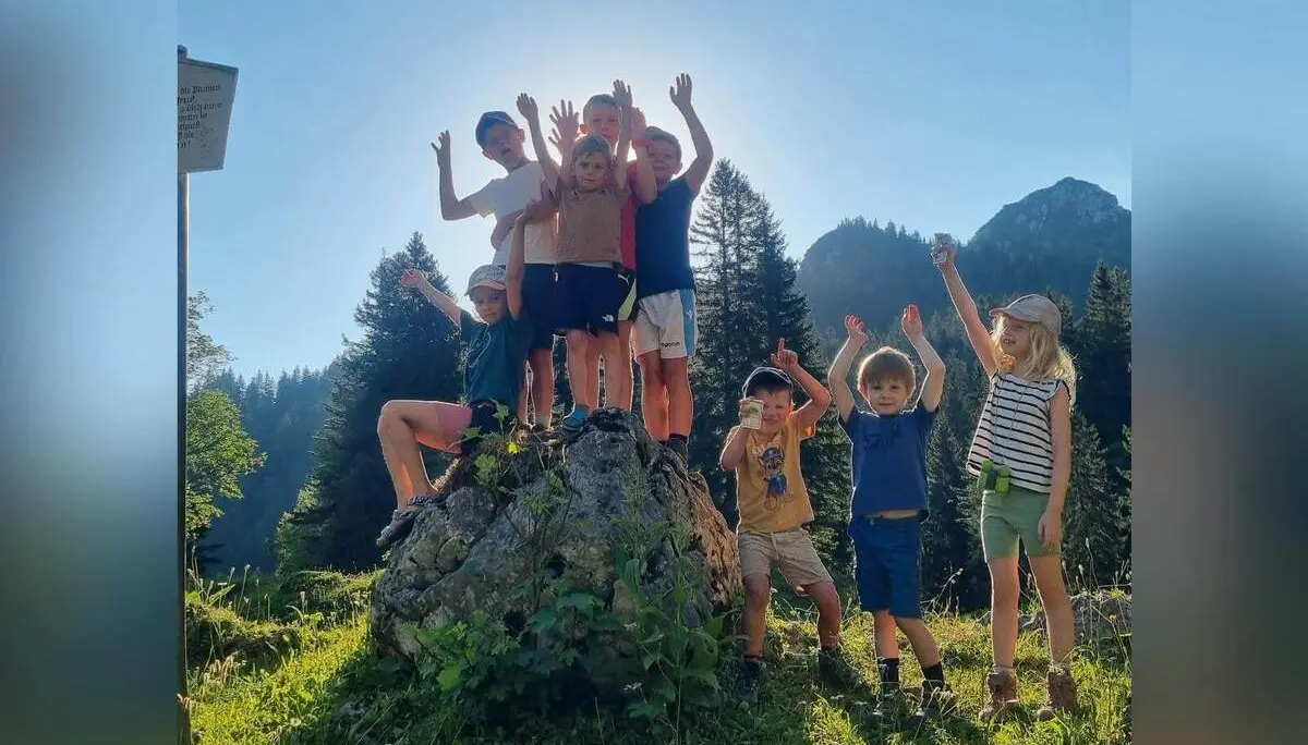 Mehrere Kinder stehen und sitzen auf einem großen Felsblock in einer Bergwiese; dahinter erheben sich bewaldete Hänge und Gipfel. | © DAV Markt Schwaben