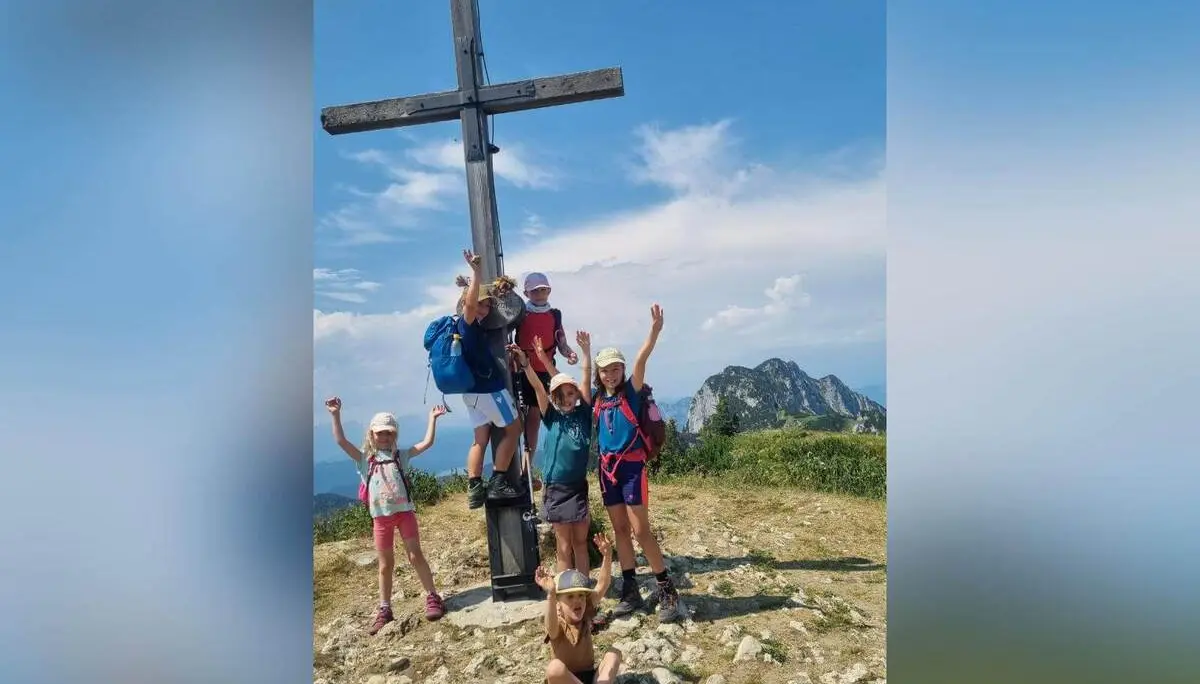 Mehrere Kinder posieren an einem Holzgipfelkreuz auf einem aussichtsreichen Berggipfel; im Hintergrund sind weitere Berge zu sehen. | © DAV Markt Schwaben