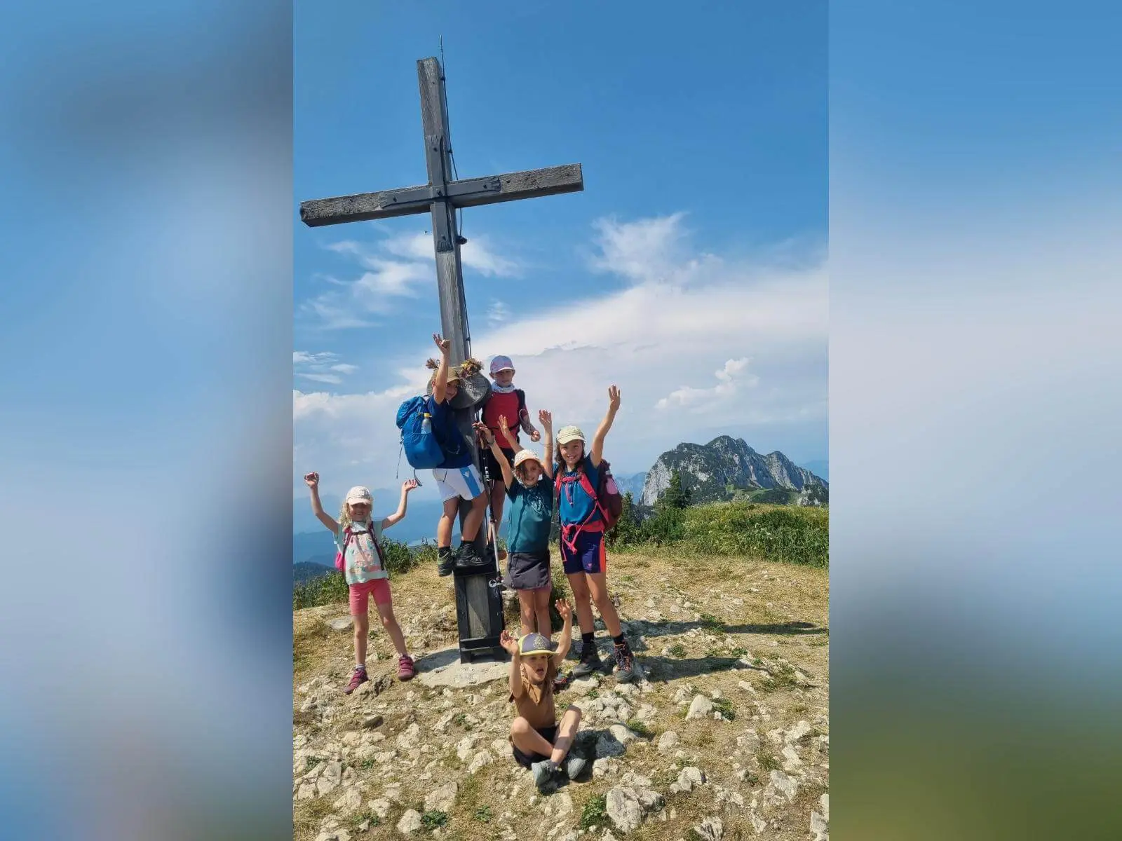 Mehrere Kinder posieren an einem Holzgipfelkreuz auf einem aussichtsreichen Berggipfel; im Hintergrund sind weitere Berge zu sehen. | © DAV Markt Schwaben