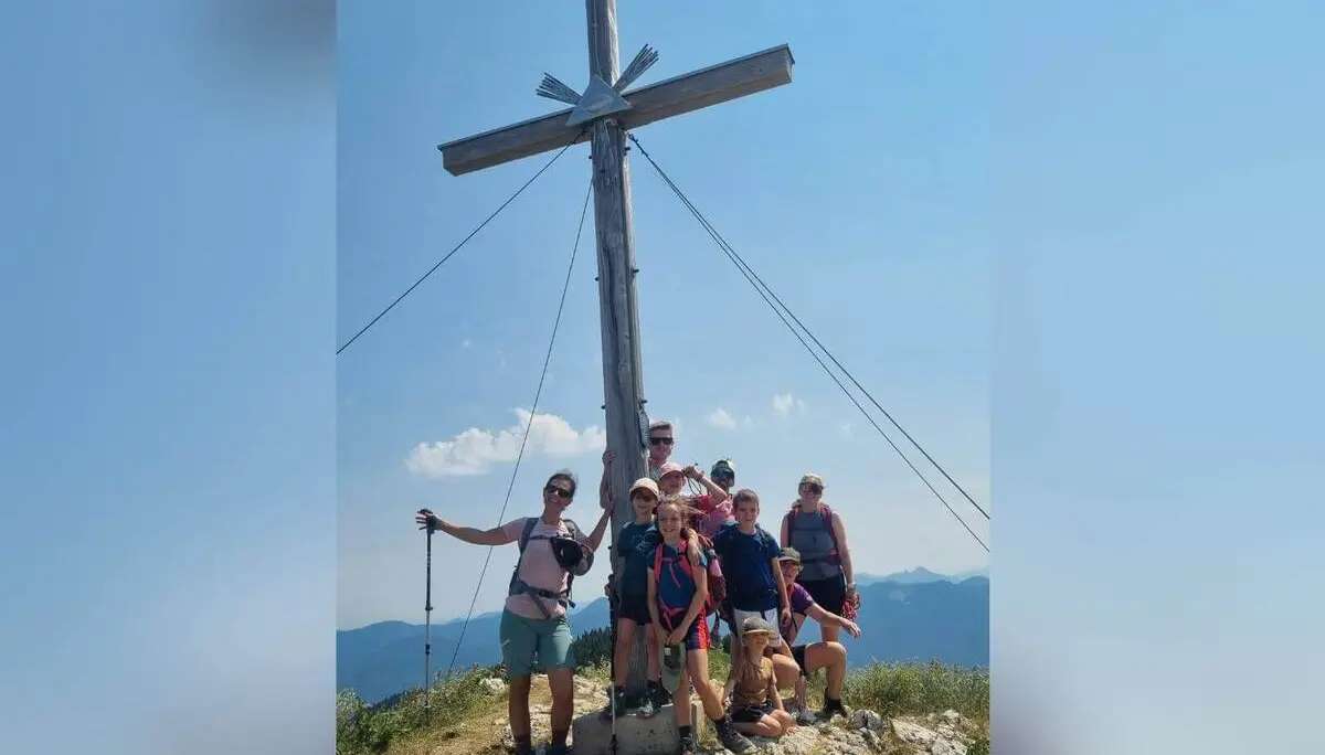 Wandergruppe mit Kindern und Erwachsenen steht unter einem großen Holzgipfelkreuz auf felsigem Berggipfel mit Fernblick. | © DAV Markt Schwaben