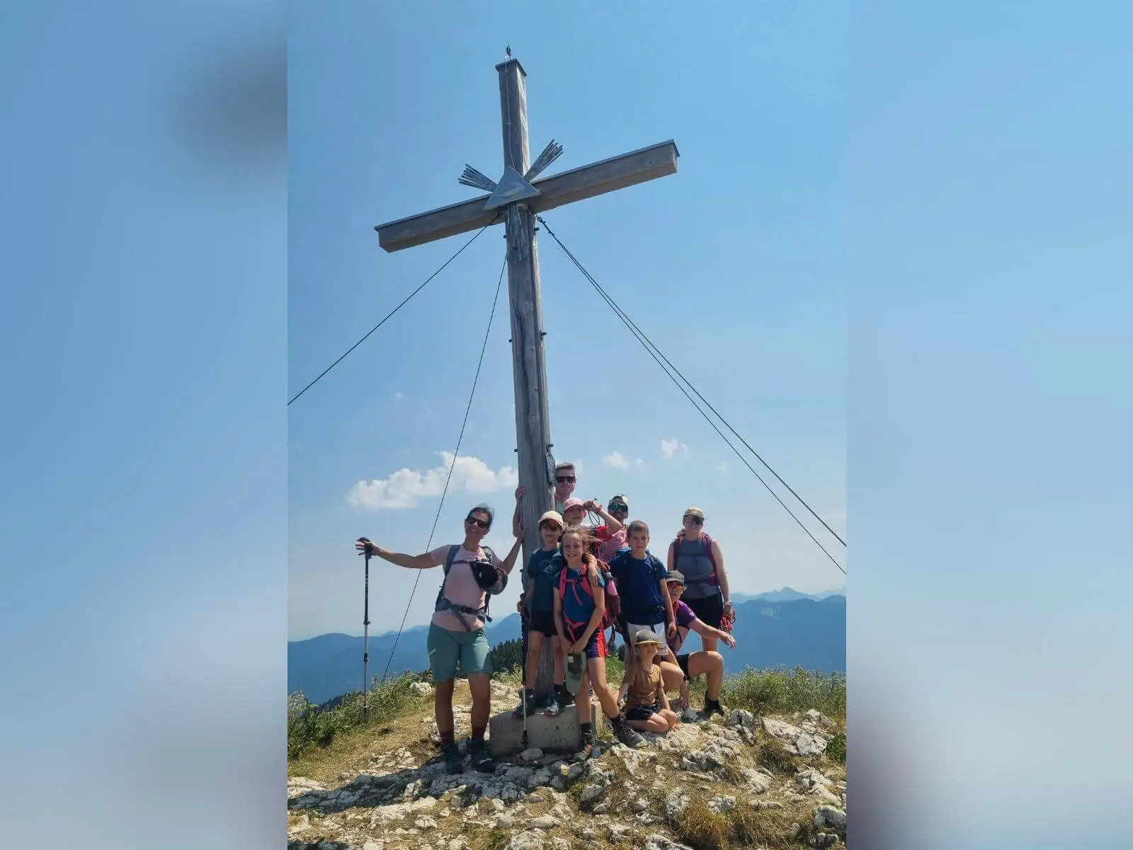 Wandergruppe mit Kindern und Erwachsenen steht unter einem großen Holzgipfelkreuz auf felsigem Berggipfel mit Fernblick. | © DAV Markt Schwaben