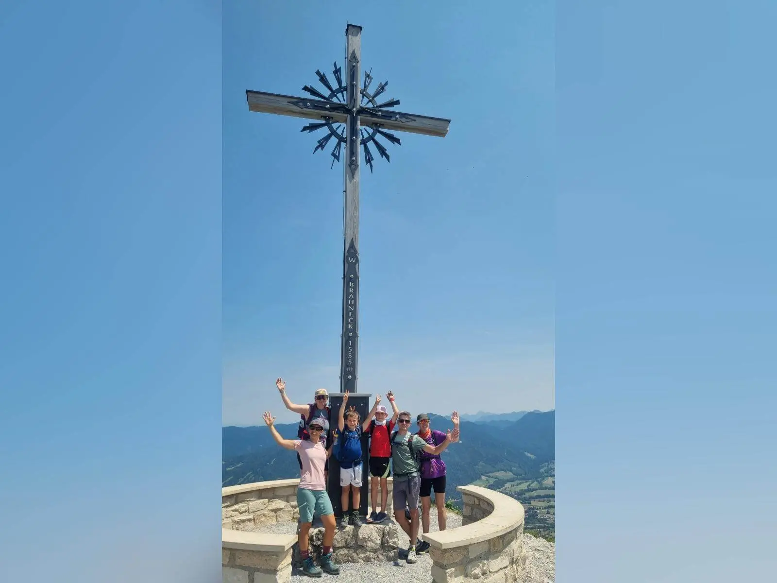Kleine Wandergruppe steht am Brauneck-Gipfelkreuz auf 1555 Metern; dahinter reichen die Blicke über Täler und Bergketten. | © DAV Markt Schwaben