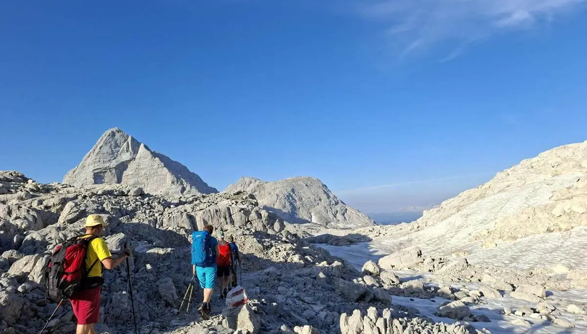 Mehrere Wandernde durchqueren eine helle Karstlandschaft mit Schneefeldern. Markante Gipfel ragen unter blauem Himmel empor. | © DAV Markt Schwaben/Alfred Quiel